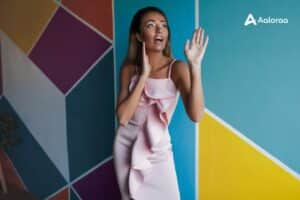 Woman in a pale pink ruffle dress stands near a colorful geometric mural, hands raised and surprised moment captured.