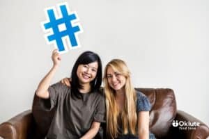 Two women sitting on a brown leather couch smiling, one holding a large blue hashtag prop.
